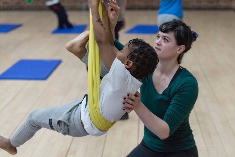 A Scarabeus tutor helps a young boy move his above him as he balances in a yellow sling.