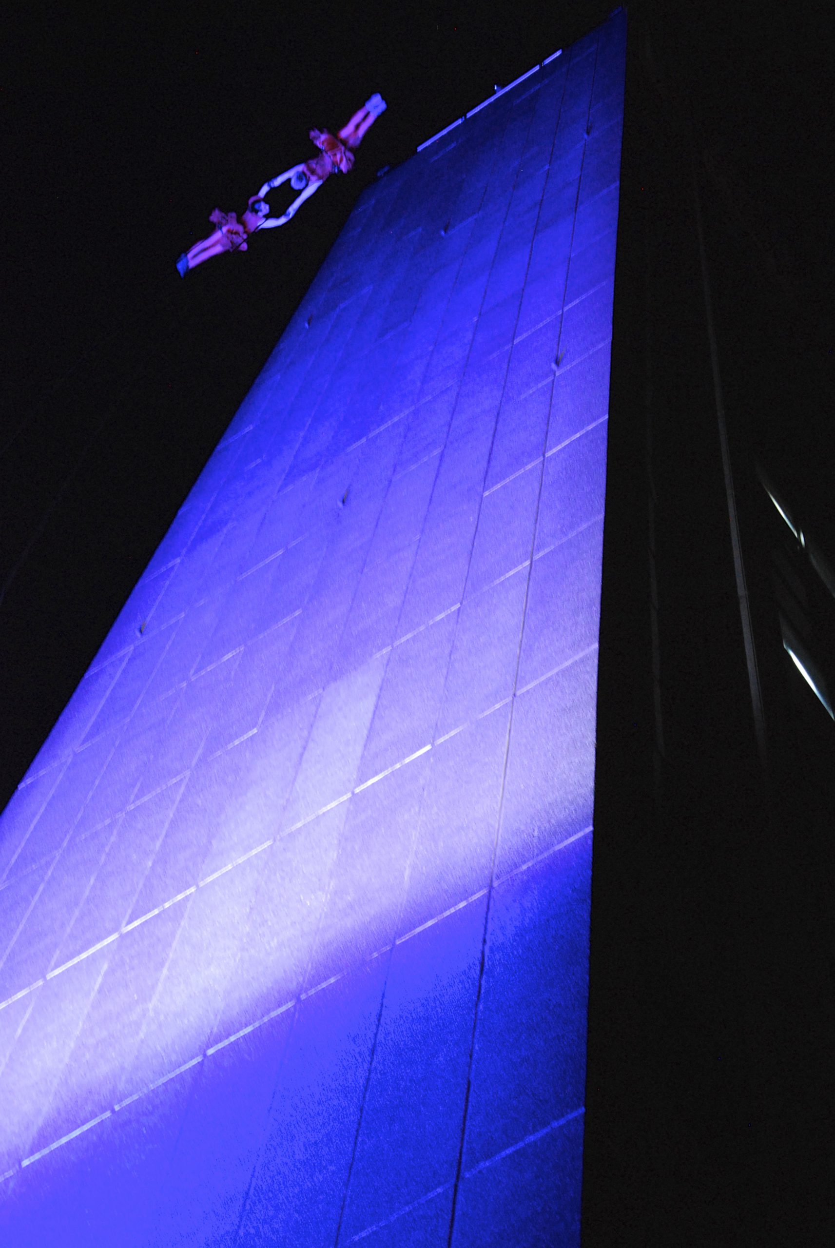 Two performers suspended looking as though they are falling from the top of a tall building spotlighted in blue