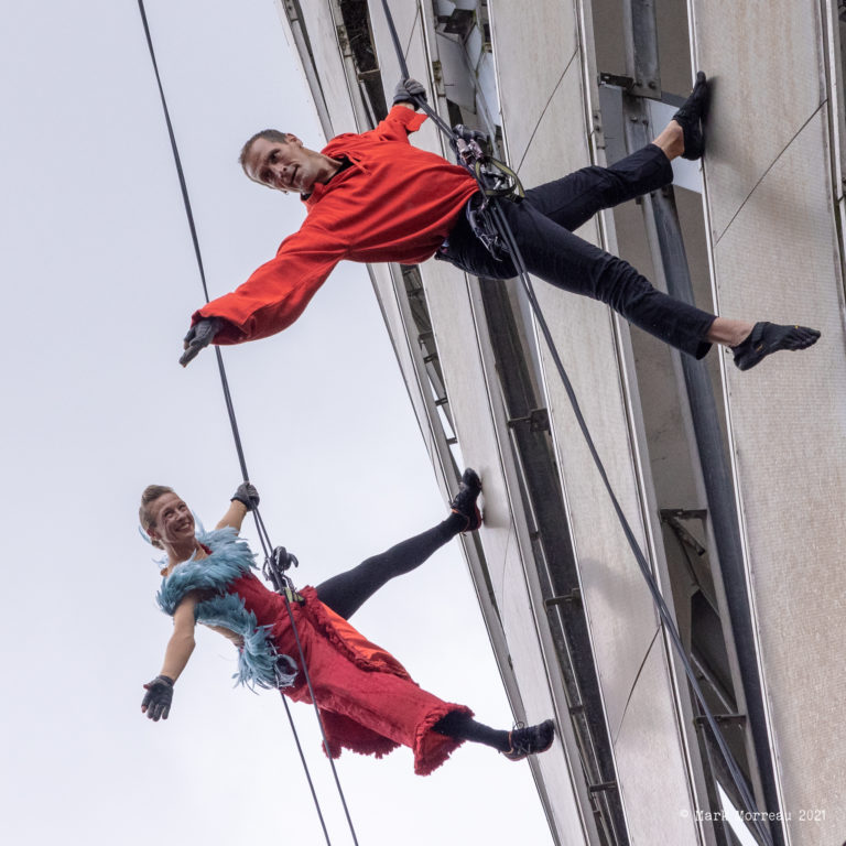 Two aerial performers suspended on ropes on the side of a building, reaching out into the air.