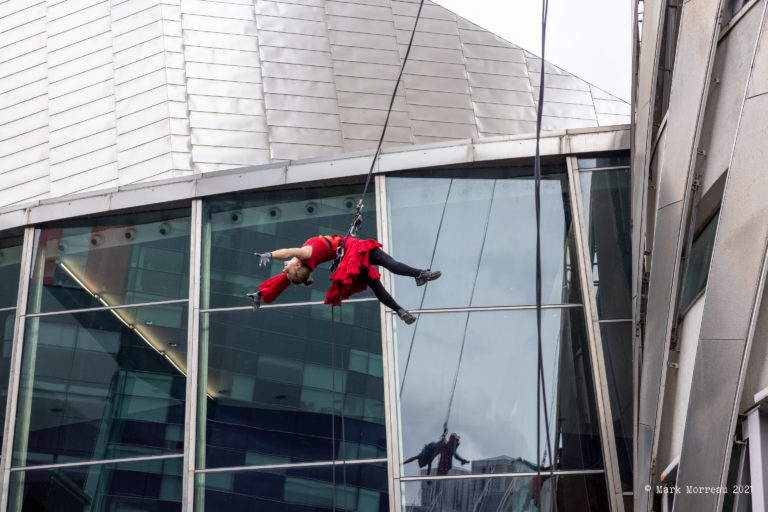 One aerial performers jumping from building with arms and legs extended, secured by harness and rope.