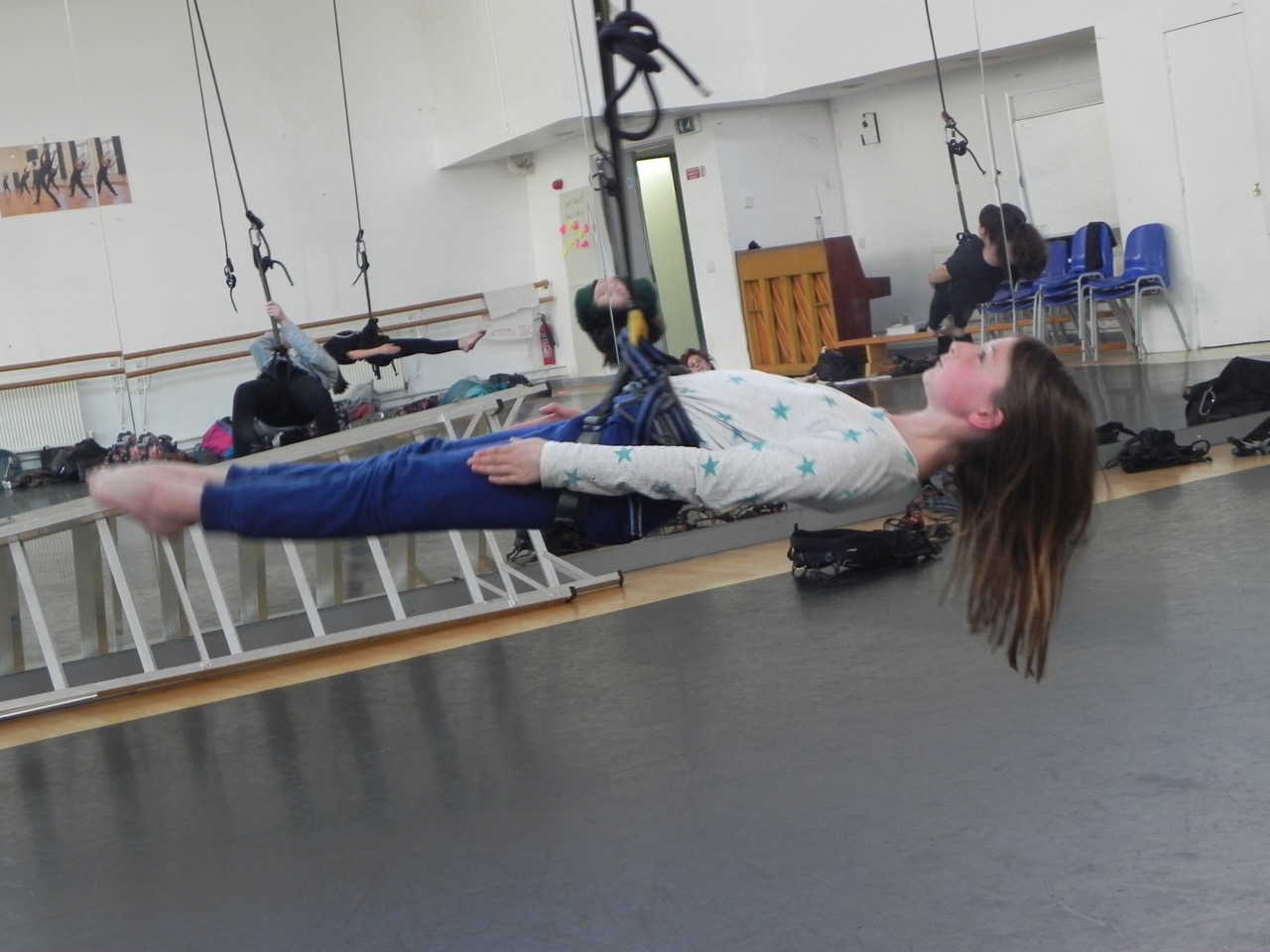 Young girl with straight plank body suspended in the air by a harness, other students reflected in the mirror behind.