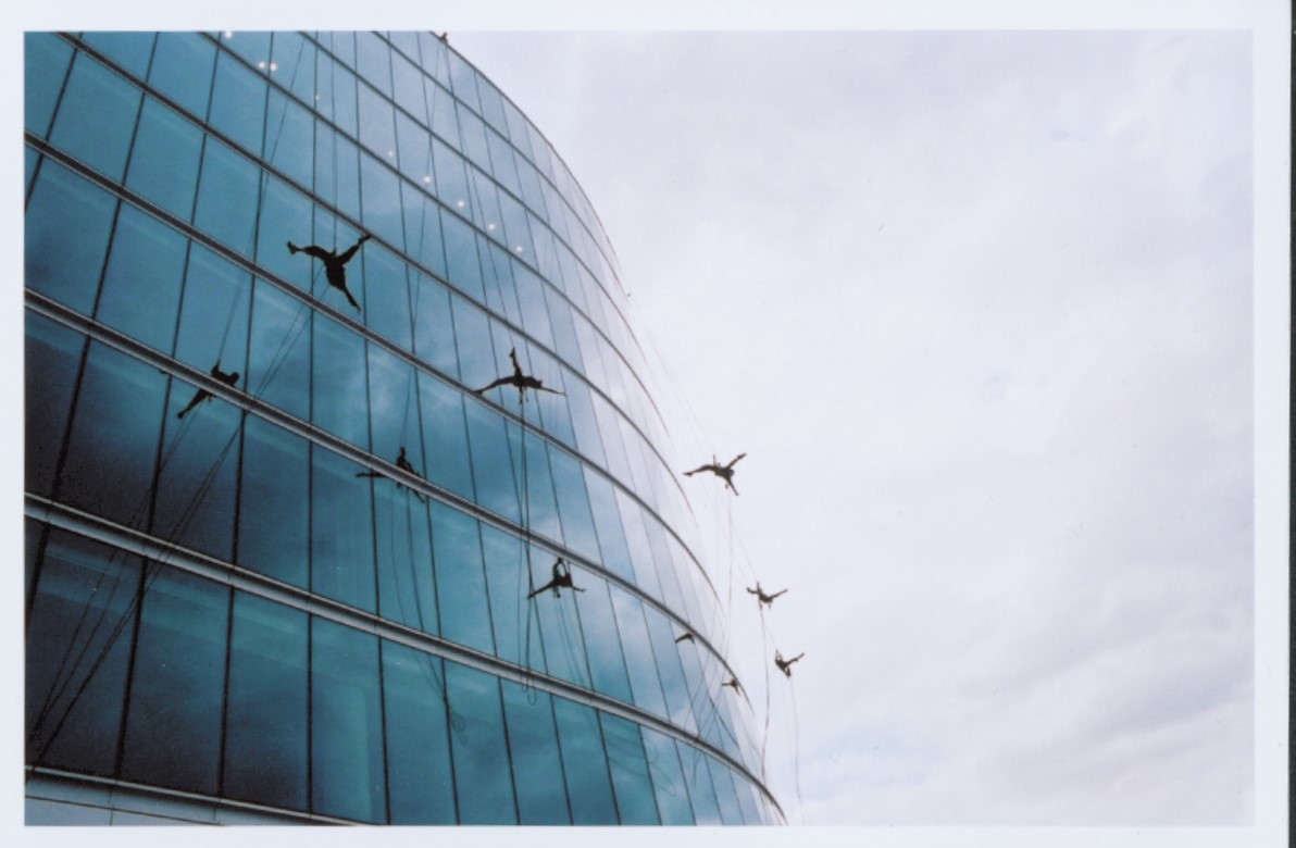 Silhouettes of 5 performers jumping from wall of glass windows of large building, suspended on ropes and reflected in the blue glass.