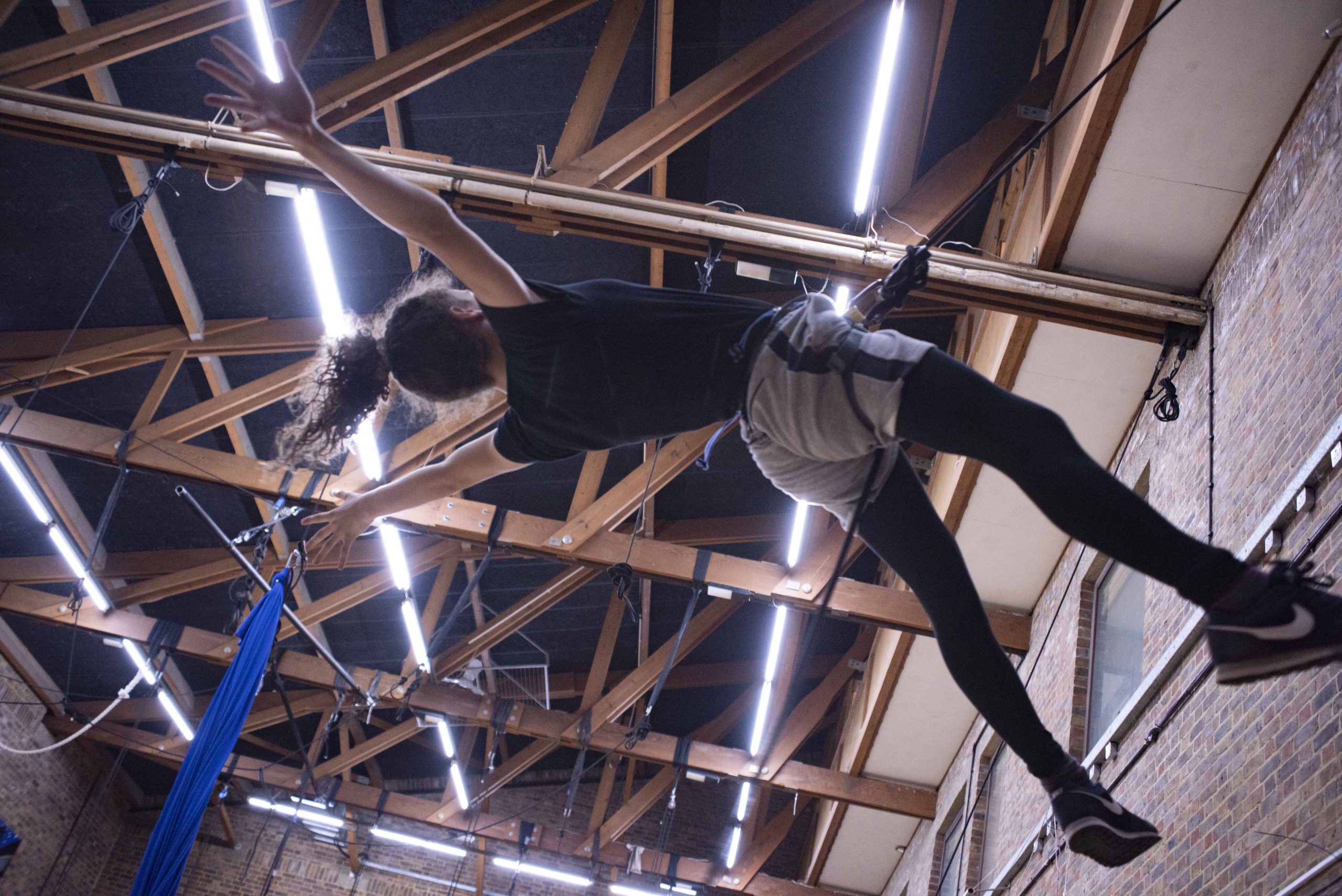 Girl with ponytail suspended with a harness from the rafters, arms open wide.