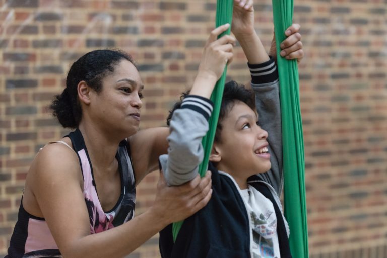 A mother supports the arms of her smiling son as he practices a move of a green cocoon.