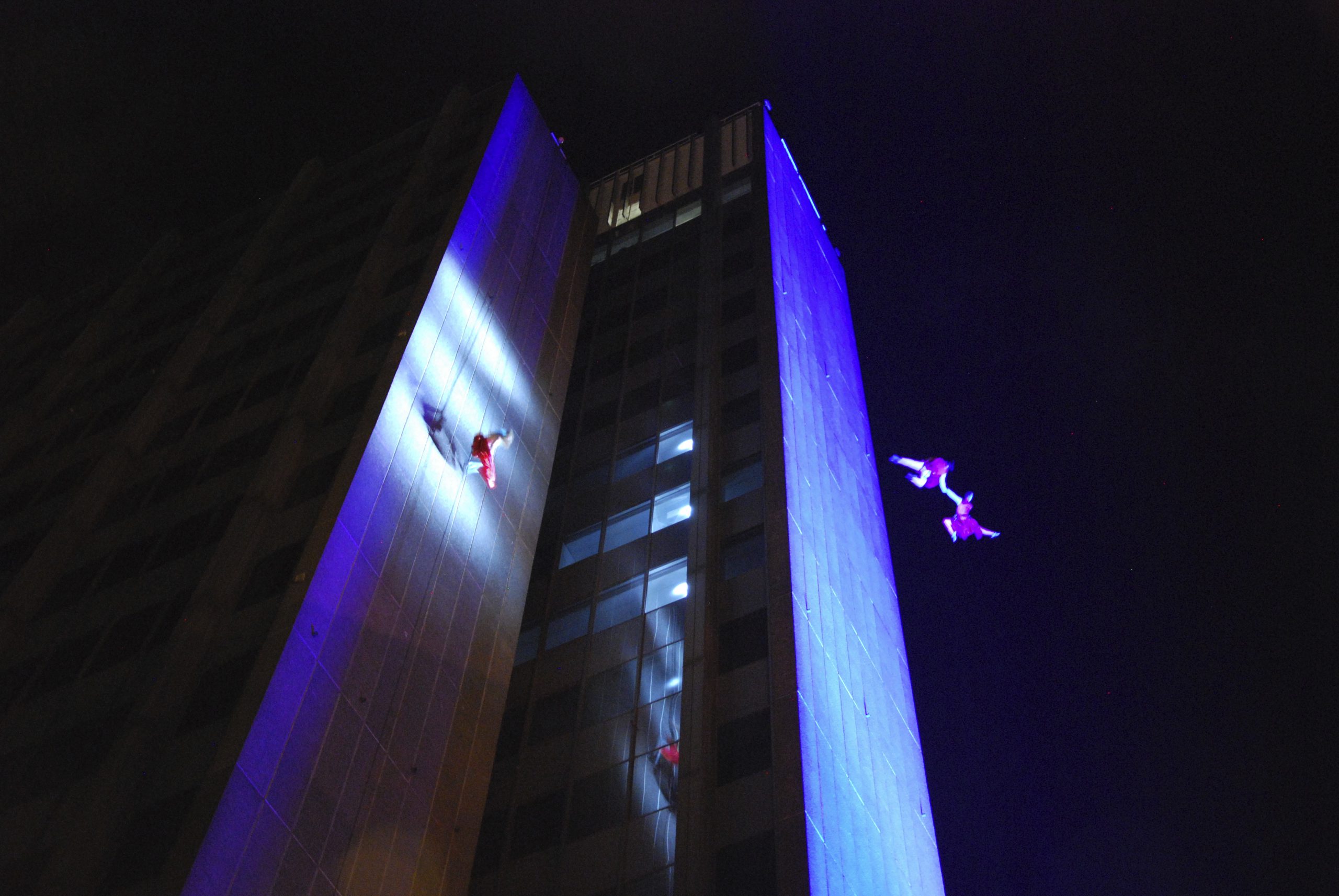 Three performers suspended from tall building (archway tower) spotlighted in blue at night