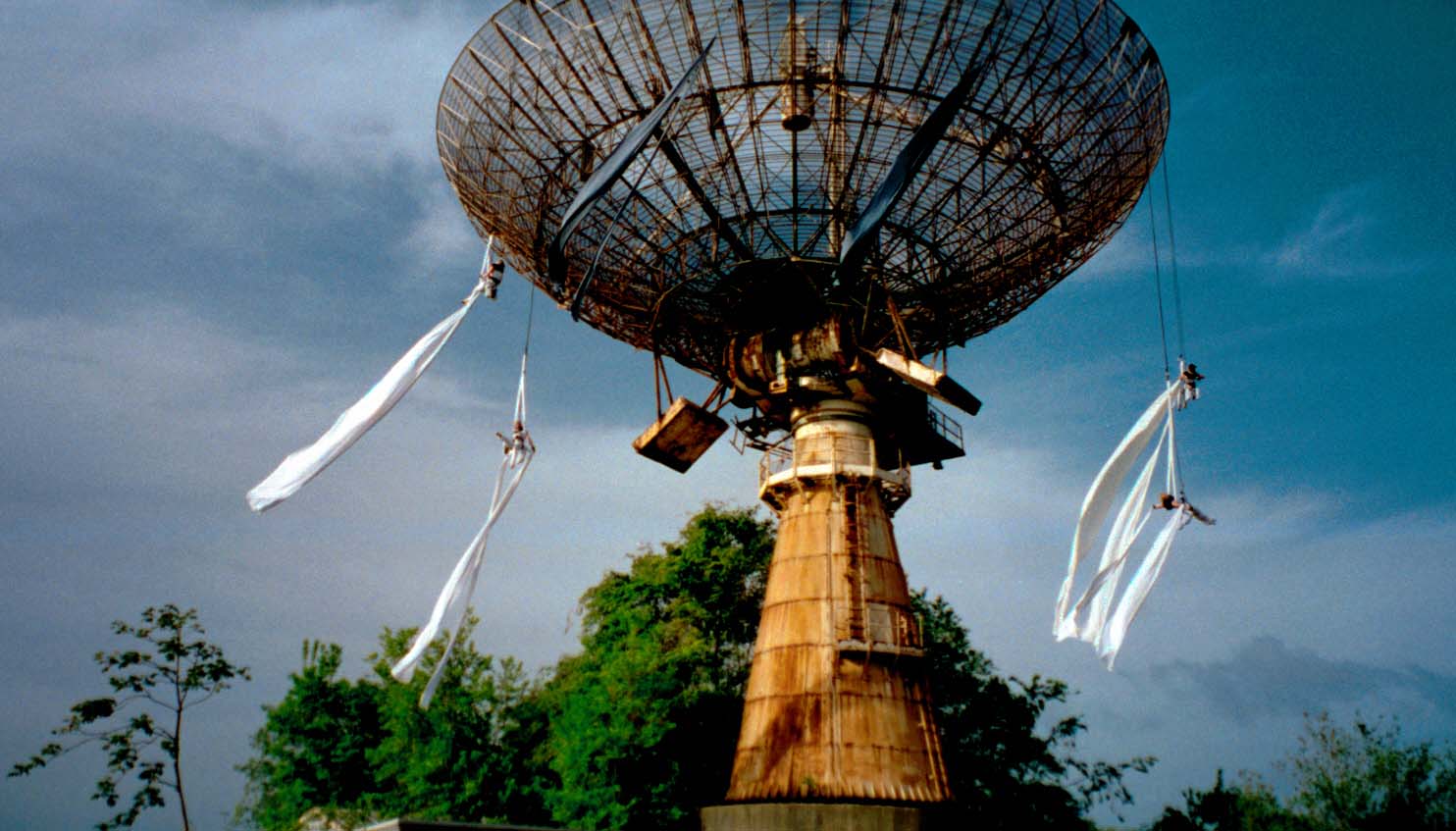Performers hanging from large satellite dish with trailing white fabric.