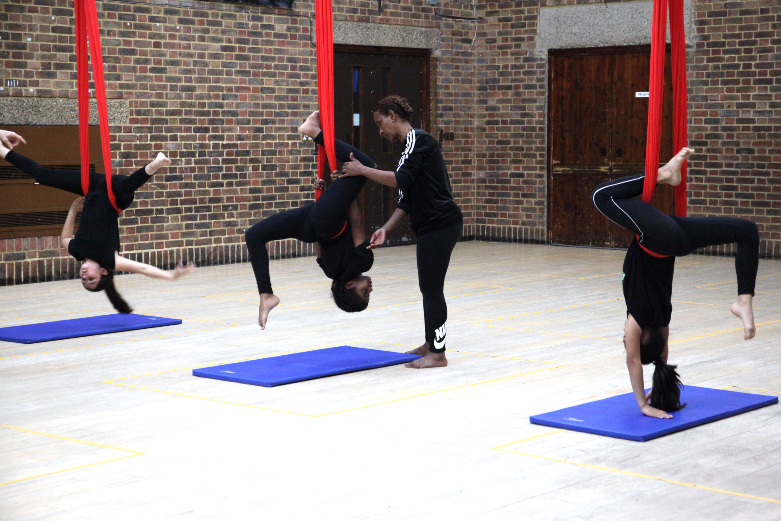 Three children are upside-down, practicing aerial moves, suspended on red silk they are each in a different pose, one with their legs and arms spread like a star, the next kicking their legs behind them with help from their mother, and the final in a similar position, with her arms touching the blue mat beneath her.