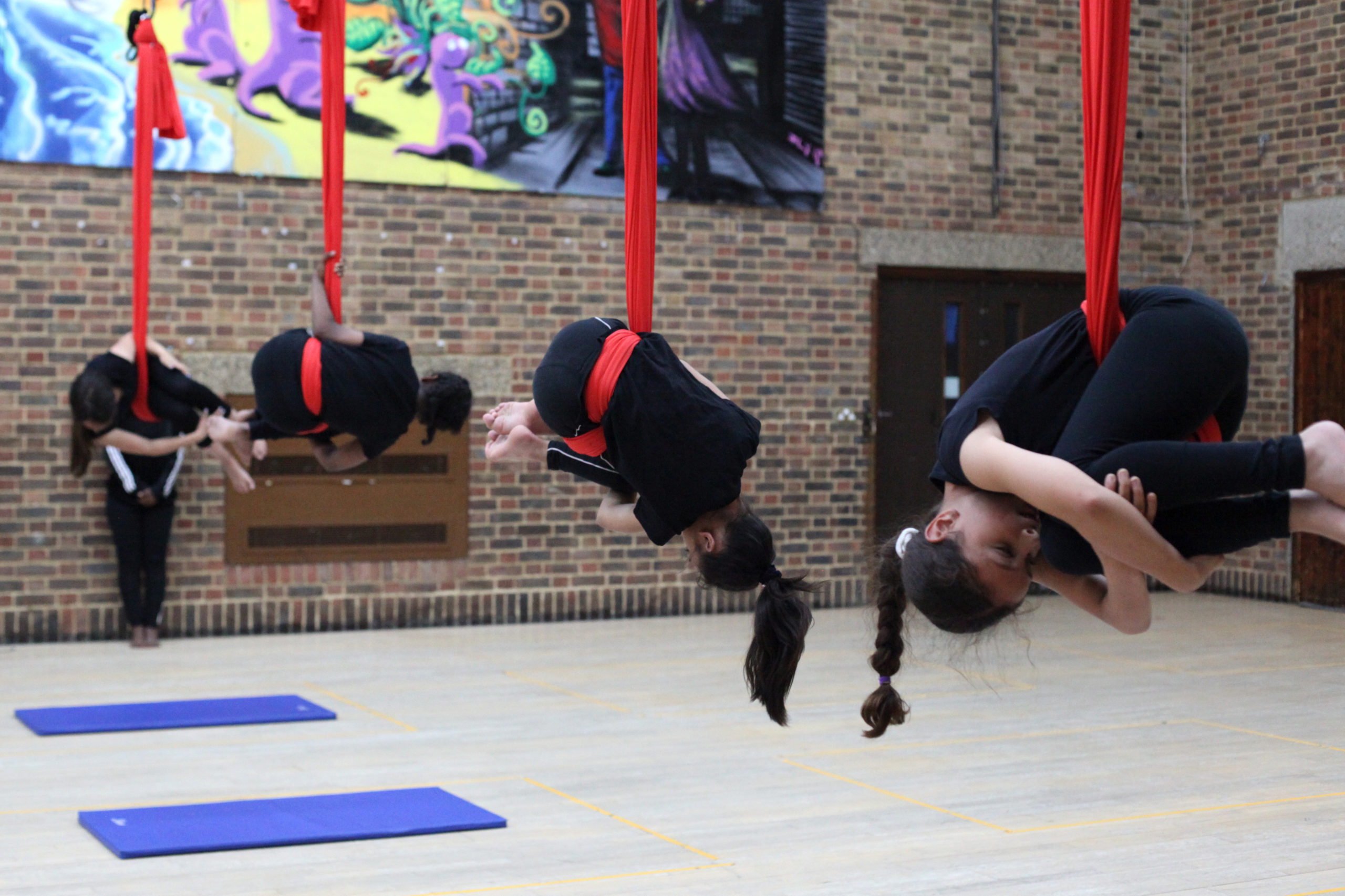 In a line, four children are wrapped in a band of bold red silk, suspended from the ceiling, they curl into a ball shape.