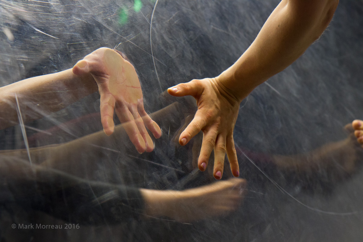 Hands are pressed, palm to palm, with a perspex screen between them. There are reflections, lights spots and scratches covering the surface.