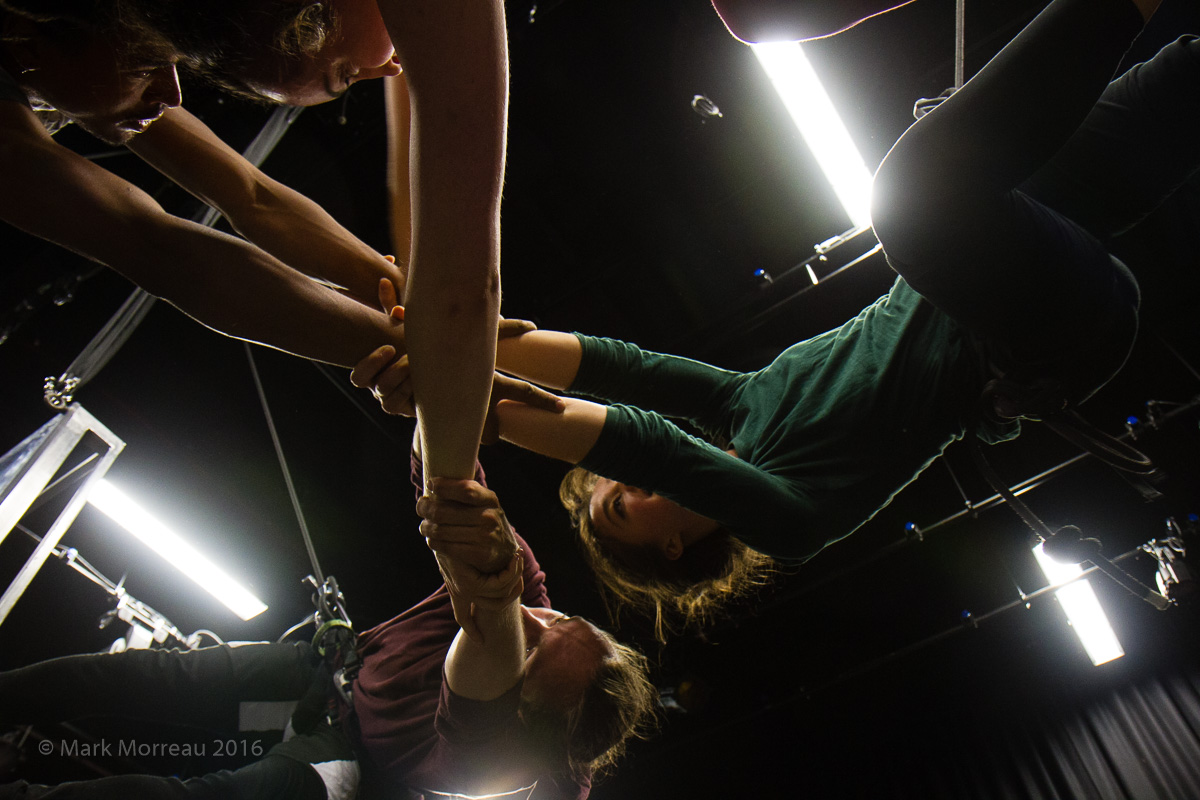 Four performers are embracing arms in pairs. They cross each other so their arms form a X, lights shine above them as they smile.