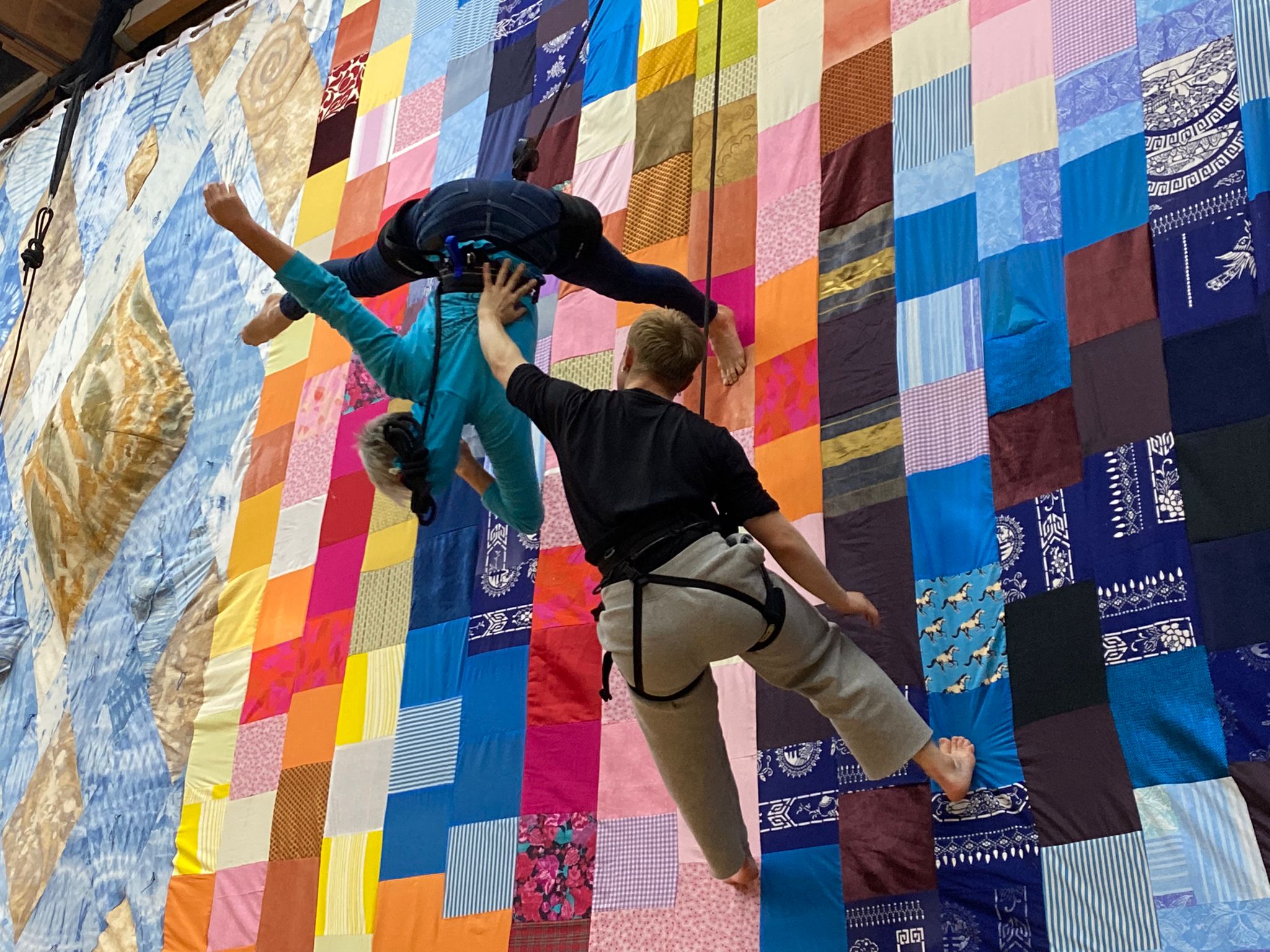 Two aerialists practice movements suspended in the air, on top of a colourful patchwork quilt.